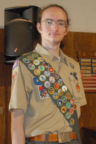 Juni stands on a stage in their full Scout uniform, displaying an impressive collection of merit badges