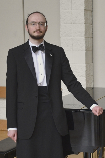 Juni wears a tuxedo and stands next to a piano at their Senior Recital