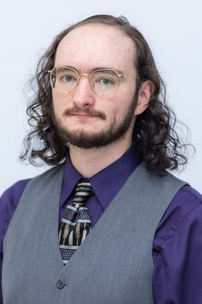 Juni's professional headshot, featuring a dark purple shirt, a patterned black tie, and a grey vest