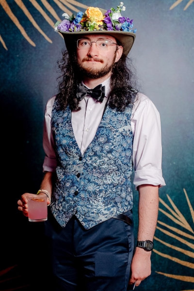 A colorful flower crown atop their iconic hat, Juni stands in a lavender shirt with black bow tie and blue floral vest, holding a pink cocktail